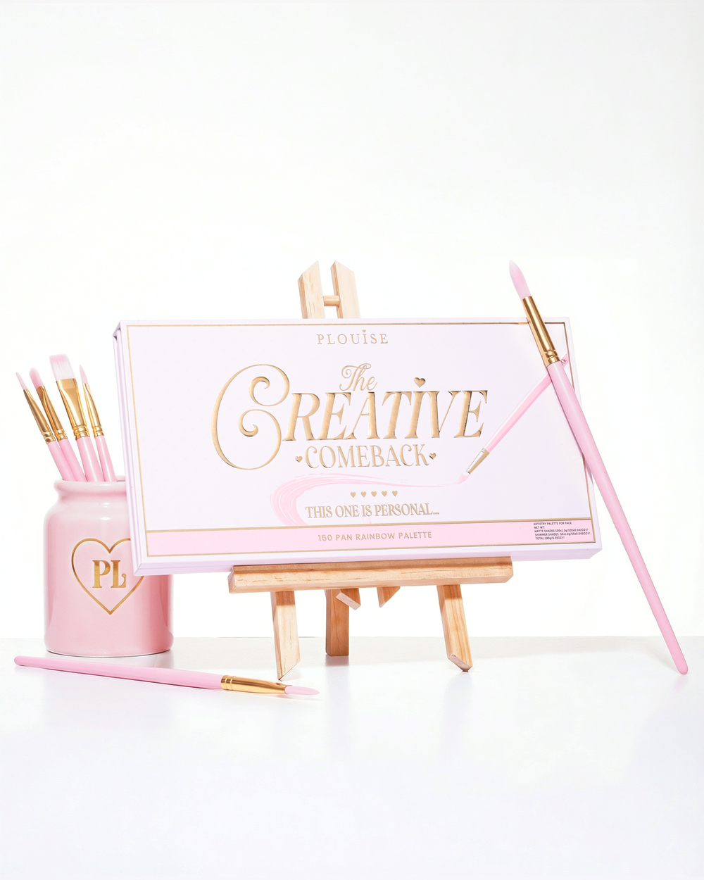 A closed pink P.Louise Creative Comeback XXXL Rainbow Eyeshadow Palette placed on an art easel surrounded by makeup brushes